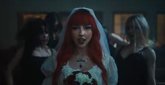 A woman with long red hair and a white veil stands in front of other people in a dimly lit setting.