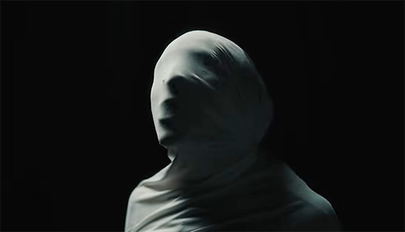 A person's head and shoulders covered in white fabric against a dark background.