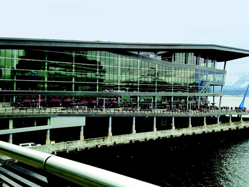 A modern multi-story glass building with a wide overhang situated along a waterfront promenade.