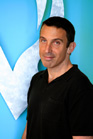 A man with short dark hair wearing a black polo shirt against a blue background.