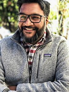 A smiling man with dark hair, glasses, and a beard wearing a grey Patagonia fleece jacket.