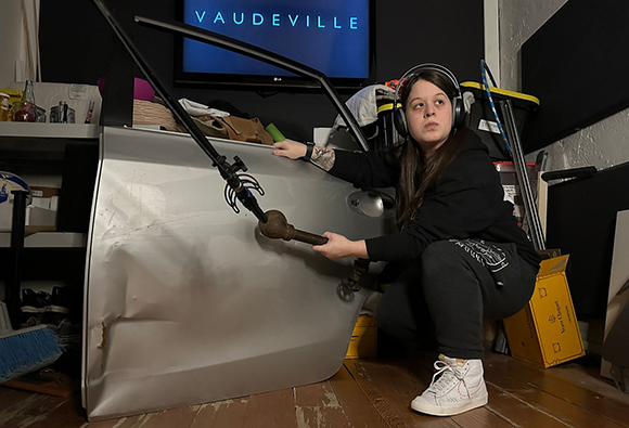 A person in a black hoodie and headphones crouches beside a dented silver car door and a microphone.