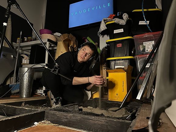 A man in black clothing crouches in a cluttered room filled with storage bins and boxes.