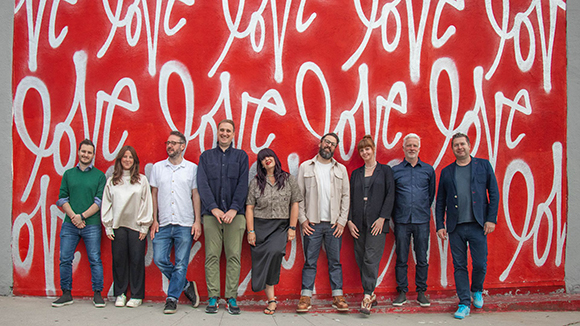 Nine diverse people standing in a line in front of a large red wall with white cursive text.