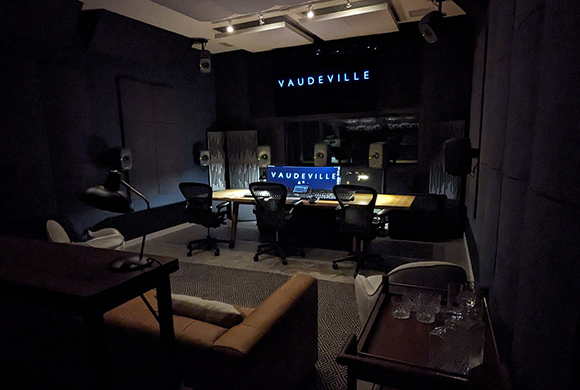 A dark recording studio featuring a mixing desk, computer monitors, and a sign reading VAUDEVILLE.