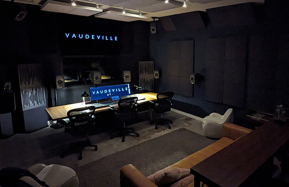 A dark recording studio with a large mixing console, computer monitors, and several ergonomic chairs.