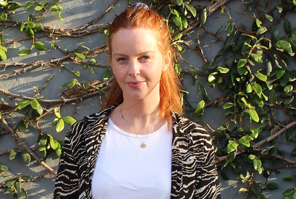 A woman with reddish hair wearing a white top and black and white patterned jacket against a green leafy wall.