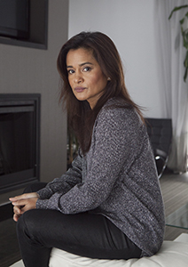 A woman with long brown hair wearing a grey sweater and black pants sits in a living room.