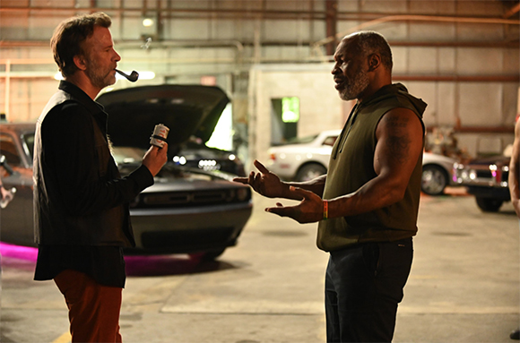 A man with a pipe talks to a muscular man in a sleeveless shirt inside a large garage with cars.