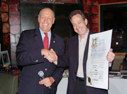 Two men in suits shake hands while one holds up a large, decorated certificate.