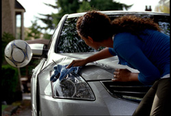 A woman in a blue shirt wipes the headlight of a silver car with a blue cloth.