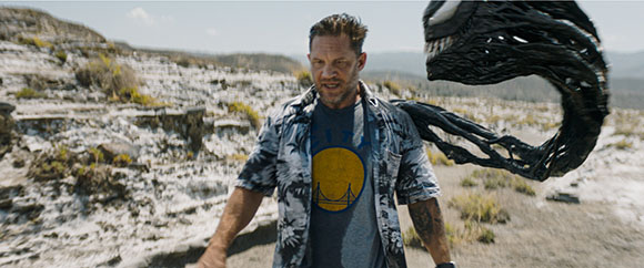 A man in a patterned shirt walks through a rocky terrain as a large black tendril looms behind him.