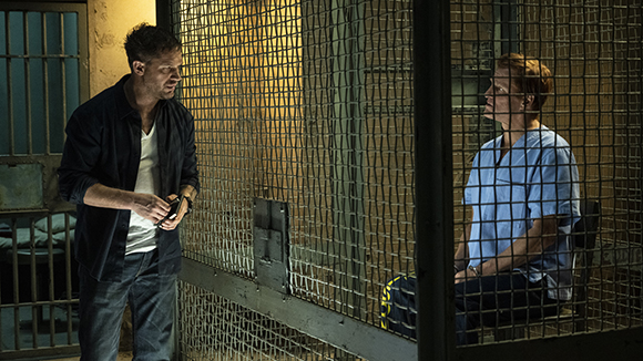 A man in a dark shirt stands outside a metal cage, talking to a person in a blue shirt sitting inside.