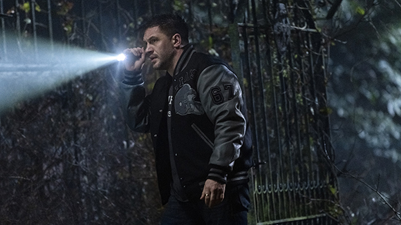 A man in a varsity jacket shines a bright flashlight through dark trees and foliage.