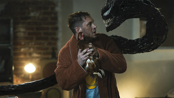 A man in a brown robe holds a brown chicken while a large, black, toothy creature curves behind him.