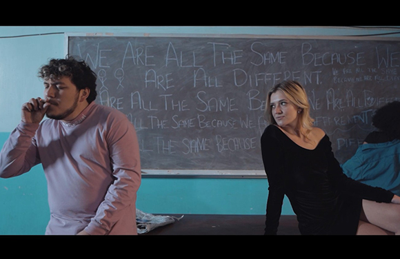 A man in a pink shirt and a woman in a black dress sit before a chalkboard with handwritten text.