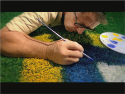 A man leans over green turf, using a thin paintbrush to apply blue paint to a yellow section.