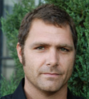 Close-up portrait of a man with short dark hair and light eyes looking forward against a green leafy background.