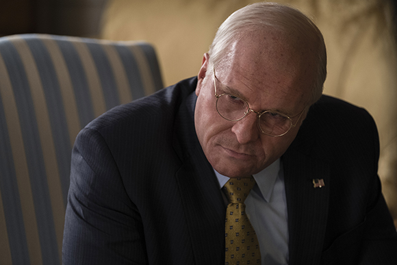 A middle-aged man in a dark suit and tie looking down with a serious expression.