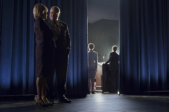 A man and woman stand in the shadows of blue curtains, watching a speaker at a podium in a bright room.