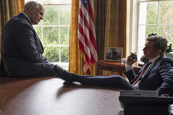 Two men in suits sit in an office; one rests his feet on a table near an American flag.