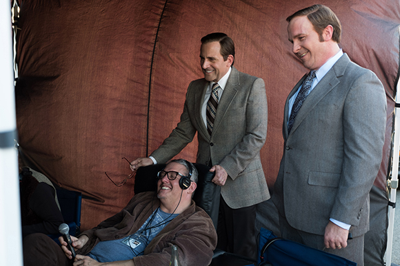 Two men in suits stand near a seated man wearing large headphones.