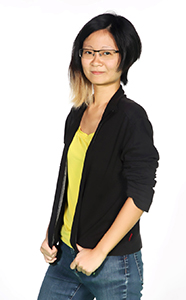 A woman with short dark hair and glasses wearing a black jacket over a yellow shirt and blue jeans.