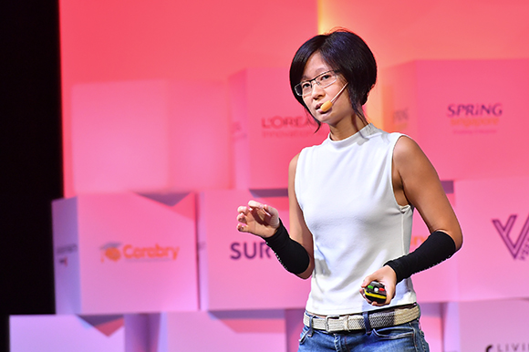 A woman in a white sleeveless top and glasses speaks into a headset microphone on a stage.