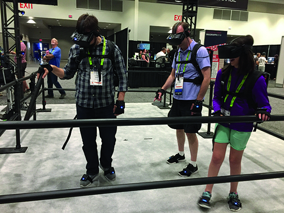 Three people wearing VR headsets and motion tracking gear walk through a fenced area at a convention.