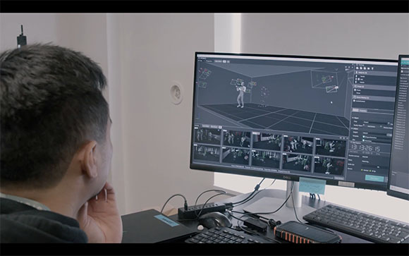 A person views a computer screen showing a 3D digital environment and multiple camera feeds.