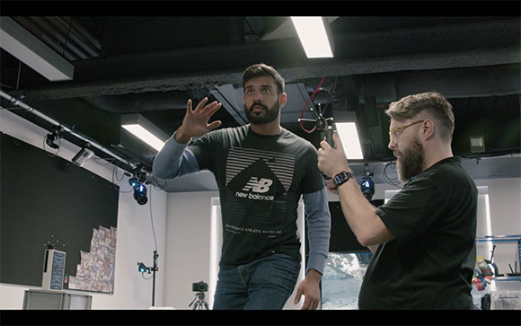 A man in a grey New Balance shirt stands in a studio while a man with glasses holds a camera toward him.