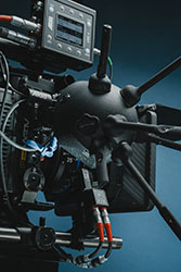 Close-up of a black professional cinema camera with various attachments and cables against a blue background.