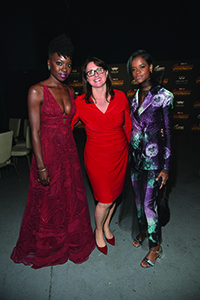 Three women posing together: one in a long red gown, one in a red dress, and one in a purple patterned suit.