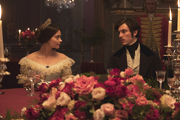 A man and woman in formal period attire sit at a candlelit dinner table behind a large bouquet of pink roses.
