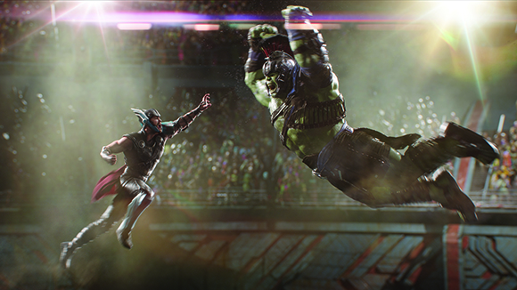 A large green muscular figure leaps through the air toward a man in dark armor in a crowded arena.