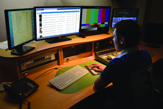 A person sits at a desk with four computer monitors, a keyboard, and various broadcasting equipment.