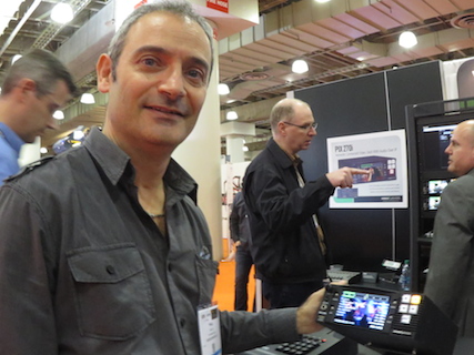 A man in a grey button-down shirt smiles while holding a small electronic monitor at an exhibition.