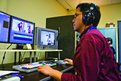 A man in a maroon shirt wearing headphones sits at a desk with multiple computer monitors and editing equipment.