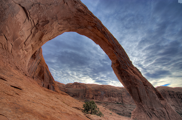 A massive red sandstone arch curves across a blue sky filled with wispy clouds.
