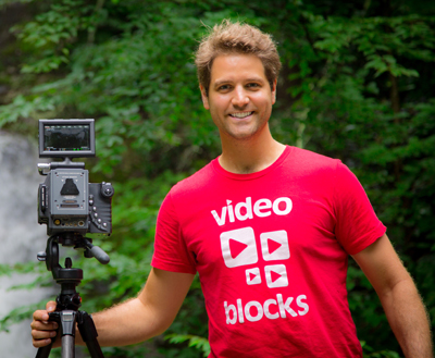 A smiling man in a red video blocks t-shirt stands next to a professional video camera on a tripod in a forest.