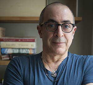 A man with glasses and a blue t-shirt sits in front of a bookshelf.