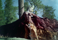 Two models in elaborate animal-themed costumes pose amidst trees in a sunlit forest.
