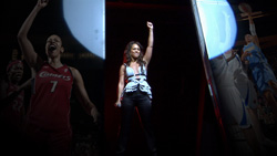 A woman raises her arm on a dark stage between large projections of basketball players.