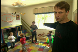 A man in a black shirt points toward three children playing in a room with a basketball hoop and colorful tiles.