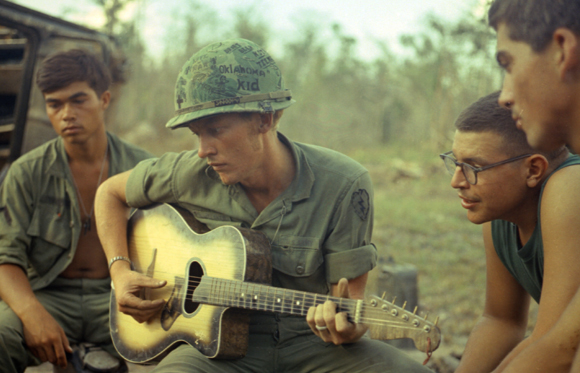 A soldier in a green uniform plays an acoustic guitar while sitting with other soldiers outdoors.