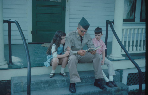 A man in a military uniform sits on porch steps reading a book to a young girl and boy.