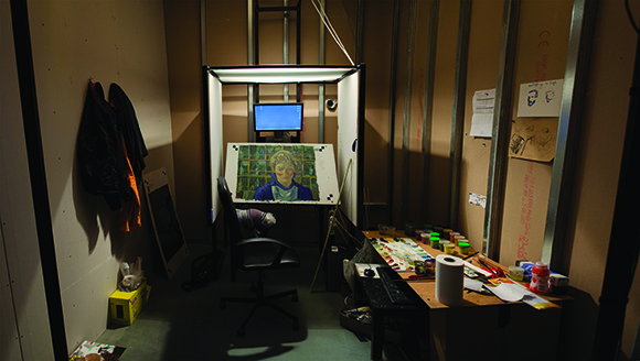A dimly lit studio with a lightbox displaying a portrait, a monitor, and a desk with art supplies.
