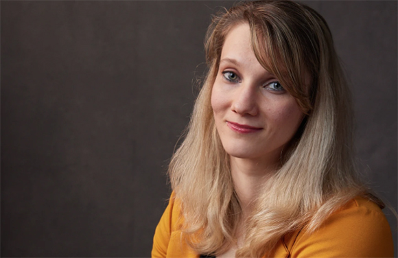 A woman with long blonde hair and blue eyes smiles slightly against a dark background.