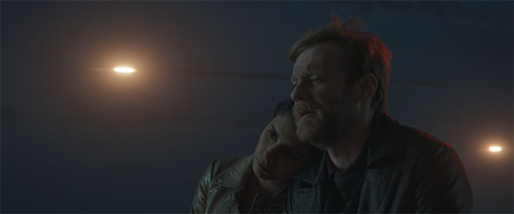 A man with a beard and a woman leaning her head on his shoulder in dim, moody lighting.