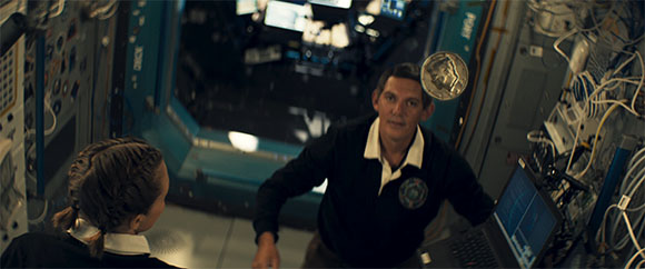 A man floats in a spacecraft near a woman and a laptop, with a coin suspended in mid-air.